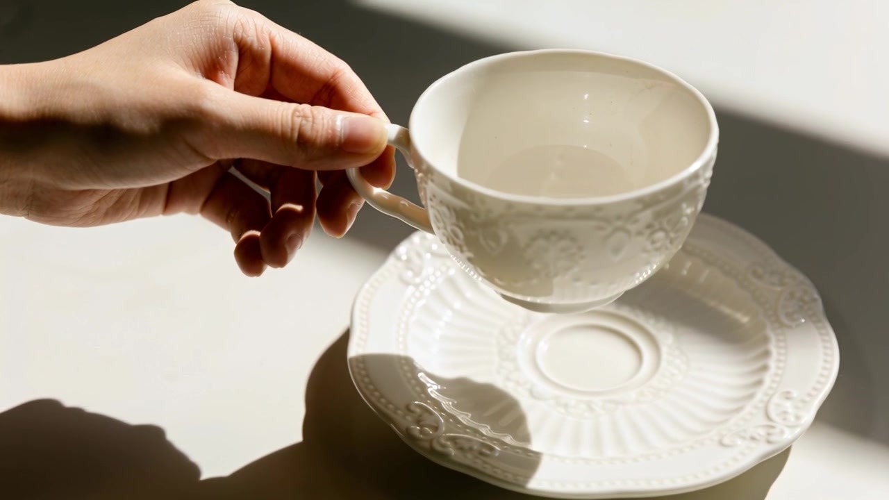 Reaching adult hand from left, grasping handle and lifting white teacup off saucer on sunlit table