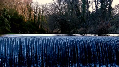 Río-Que-Fluye-Sobre-Una-Pequeña-Presa-De-Desvío-En-La-Coruña-España-Con-árboles-Forestales-Otoñales-En-El-Fondo