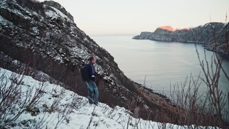 Junger-Mann,-Der-Am-Rand-Eines-Schneebedeckten-Berges-Spazieren-Geht,-Still-Steht,-Um-Die-Landschaft-Zu-Genießen,-Und-Einen-Wunderschönen-Berg--Und-Meerblick-überblickt