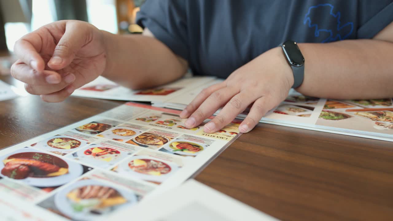 Individual points at illustrated menu, choosing dishes in well-lit Bangkok restaurant, medium close-up shot