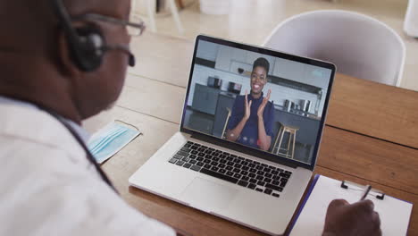 african american male doctor wearing phone headset taking notes while having a video call on laptop