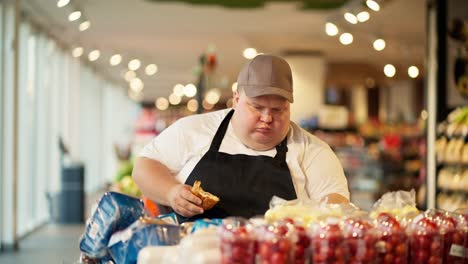 Ein-übergewichtiger-Männlicher-Supermarktangestellter-In-Einem-Weißen-T-Shirt-Und-Einer-Schwarzen-Schürze-Isst-Ein-Croissant-Und-Arrangiert-Lebensmittel-Auf-Dem