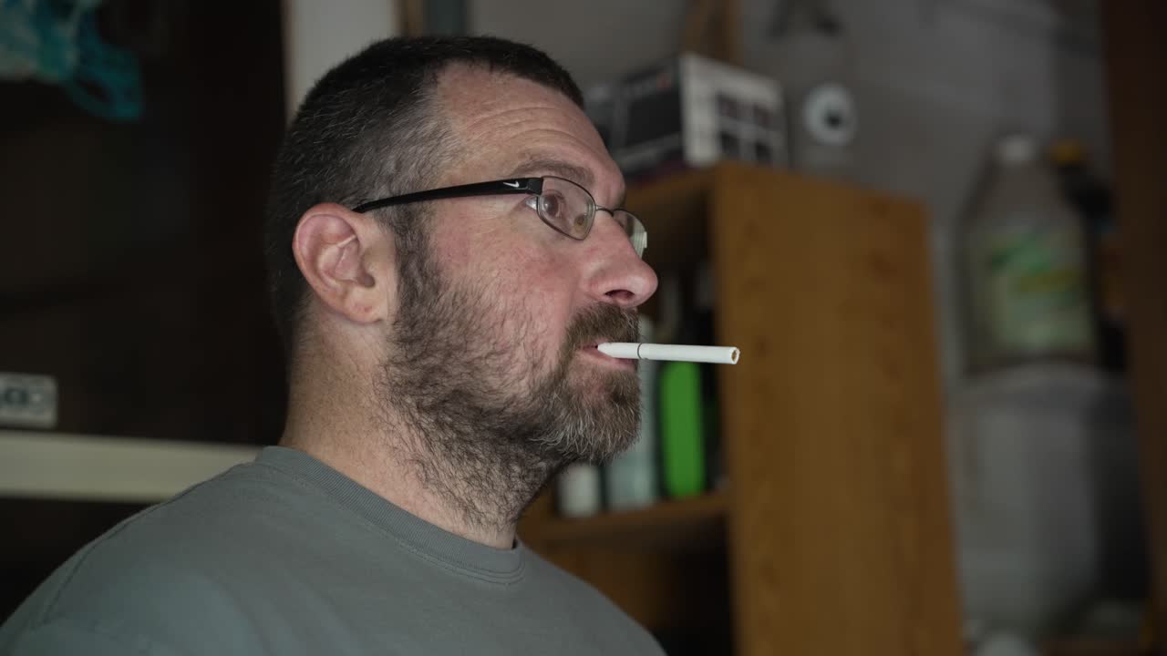 A caucasian man with glasses lights a cigarette and breathes out smoke.