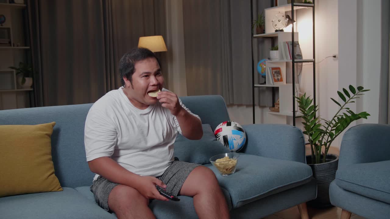 Uomo asiatico robusto con il telecomando della TV che preme un pulsante e cambia canale, seduto su un divano a casa e mangia patatine.