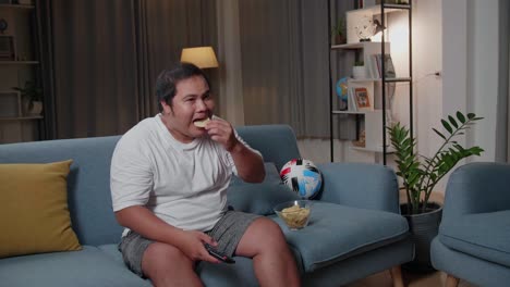 asian fat male with tv remote pressing button and changing, switch channels while sitting on a couch at home and eats potato chips