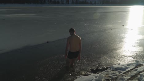 Helles-Sonnenlicht-Scheint-Auf-Die-Gefrorene-Oberfläche-Des-Bergsees,-Während-Der-Mann-Mit-Nur-Kurzen-Gängen-Ins-Wasser-Geht,-Wo-Eis-Gebrochen-Wurde