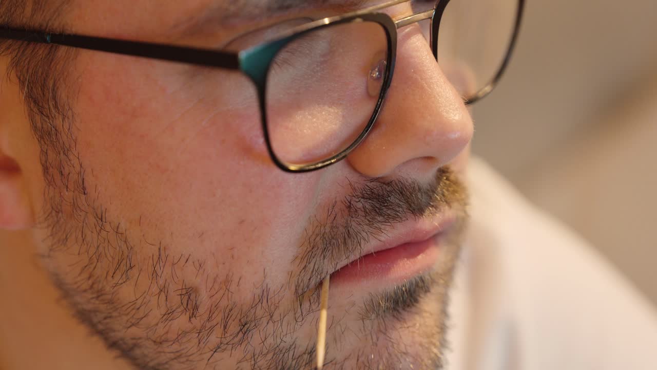 A close-up of a man’s face with a toothpick in his mouth, showing a relaxed and thoughtful expression under warm indoor lighting