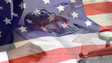 Animation-of-American-flag-waving-over-African-American-businessman-using-calculator