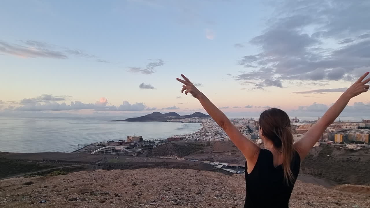 mujer admira con los brazos levantados y desde un mirador, al atardecer, la playa de las canteras y el auditorio alfredo kraus