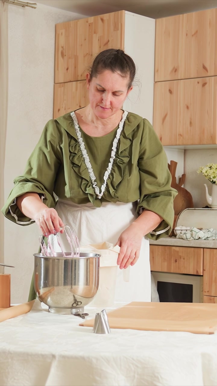 Mujer con blusa verde y delantal blanco prepara ingredientes. Luego, la mujer vierte el contenido de otro recipiente. La escena continúa con ella limpiando una batidora eléctrica bajo el agua corriente.