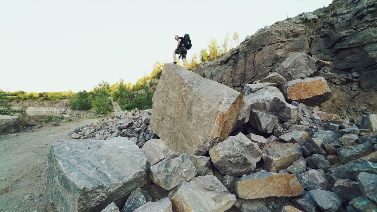 A traveler in shorts and with a backpack climbs onto a huge stone and sits on it amid a quarry with a lake in the center.