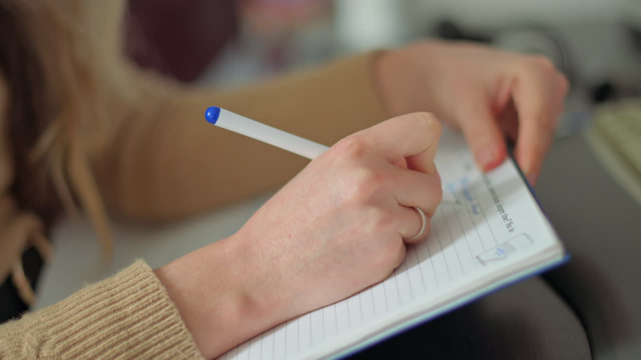 A person sits at a desk, writing notes in a notebook with a blue pen. The setting is warm and inviting, with soft lighting creating a calm atmosphere