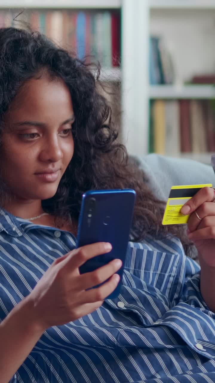 Young joy indian woman zoomer with mobile phone and credit card sits on sofa