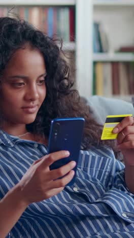 young joy indian woman zoomer with mobile phone and credit card sits on sofa