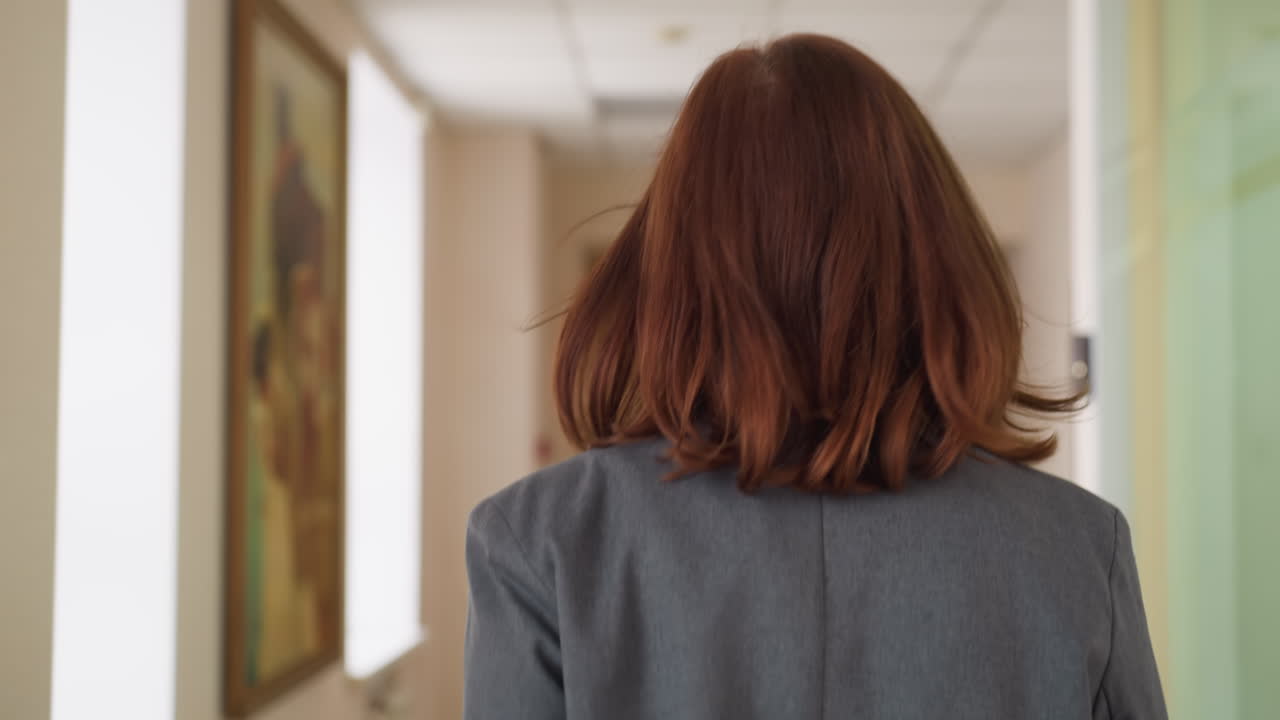 Close up back shot of woman walking purposefully down modern hallway, holding red folder, dressed in professional attire, exuding confidence and determination
