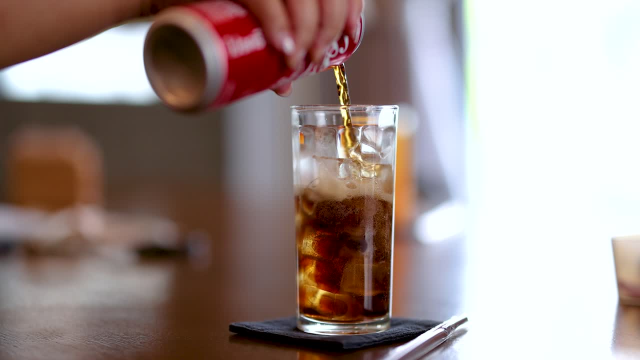 Hand pours cola into ice-filled glass, natural daylight, shallow focus, indoor dining setting, steady shot