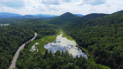 Imágenes-Aéreas-De-Una-Carretera-Que-Atraviesa-Las-Montañas-Del-Norte-Del-Estado-De-Nueva-York-Junto-A-Un-Lago-Reflectante