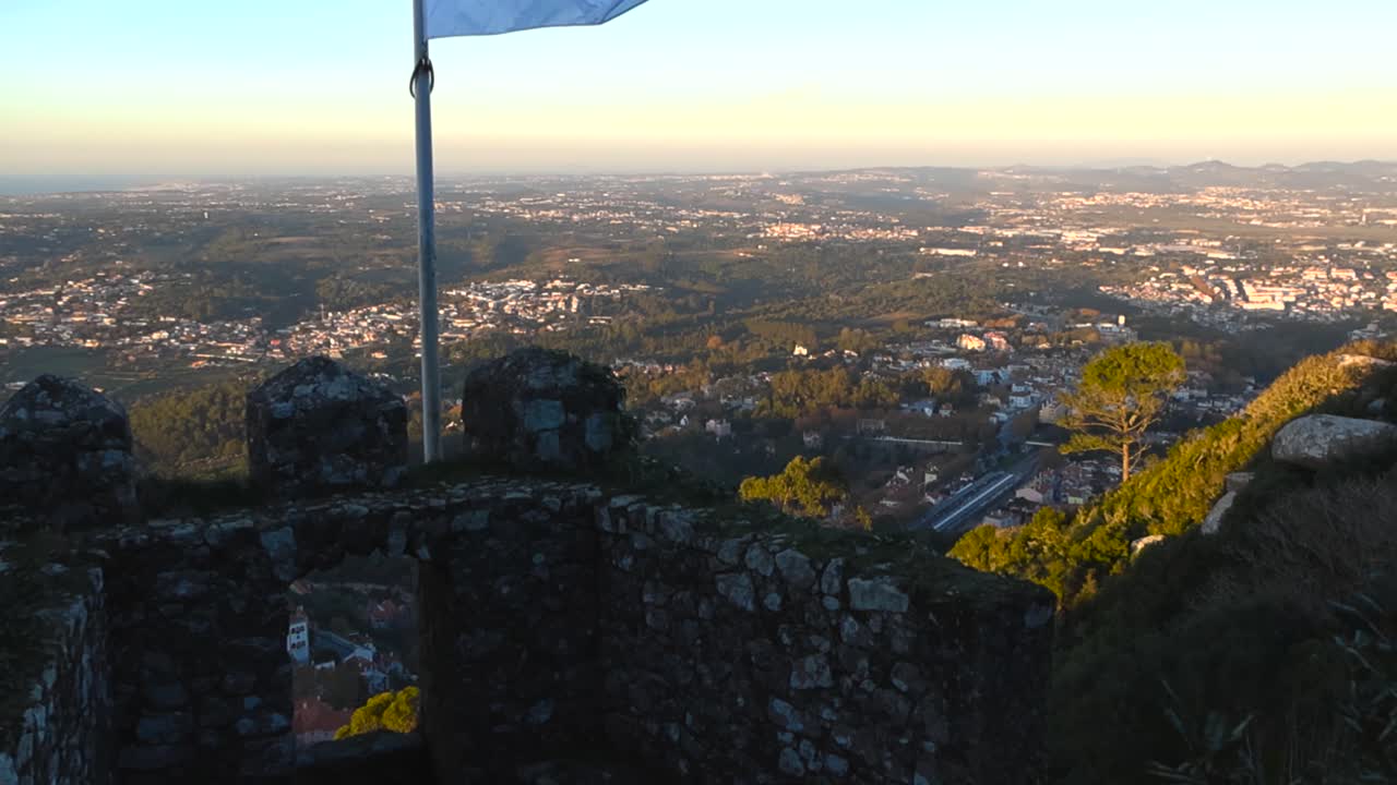 Een gouden zonsondergang verlicht het Portugese landschap beneden, gezien vanaf de bemoste muren van het Moorse kasteel, met een wapperende vlag boven glooiende heuvels en afgelegen steden