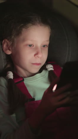 una vista de cerca de un niño que opera un teléfono celular en un coche por la noche, el niño está usando un abrigado rosa y una camisa verde claro, centrado en la pantalla del teléfono celular