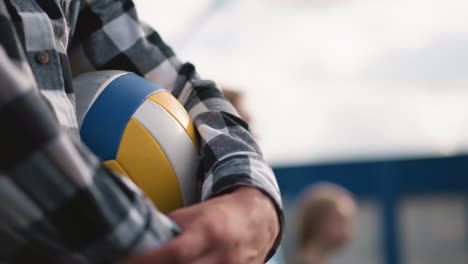 primer plano de una persona con camisa a cuadros sosteniendo una pelota de voleibol con fondo borroso con dos personas de pie cerca, el enfoque suave en el fondo crea profundidad, enfatizando los deportes y el trabajo en equipo