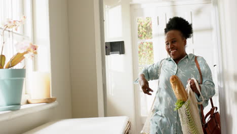 Feliz-Mujer-Mayor-Afroamericana-Entrando-A-Casa-Con-Compras-En-Una-Habitación-Soleada,-Cámara-Lenta