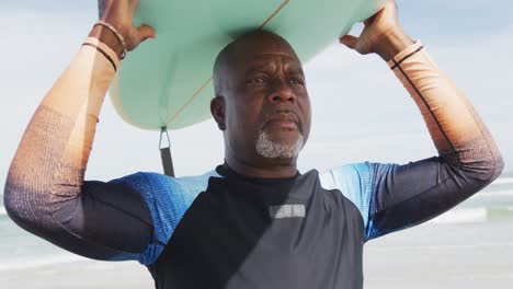 hombre mayor afroamericano caminando por una playa sosteniendo una tabla de surf en su cabeza