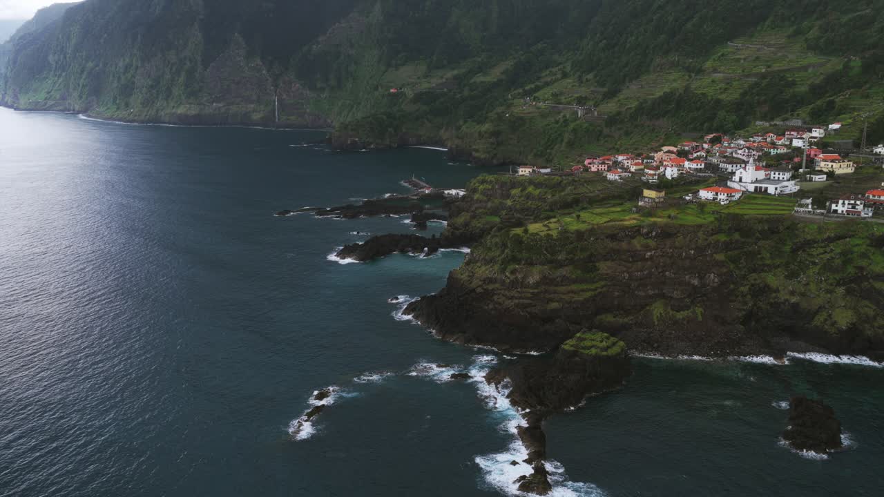 Vila costeira do Seixal na costa rochosa, ondas do Oceano Atlântico, falésias verdes íngremes e cachoeira distante, Madeira, Portugal. Vista aérea de drone