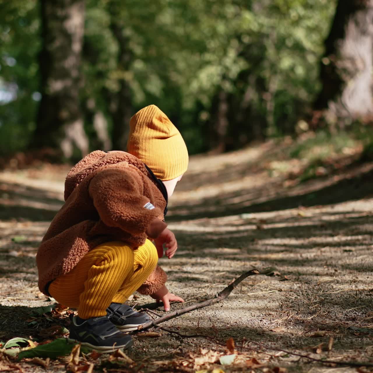 Liten småbarn sitter på huk i gränden i parken. Barnet rör vid pinnen och ställer sig sedan upp och går iväg