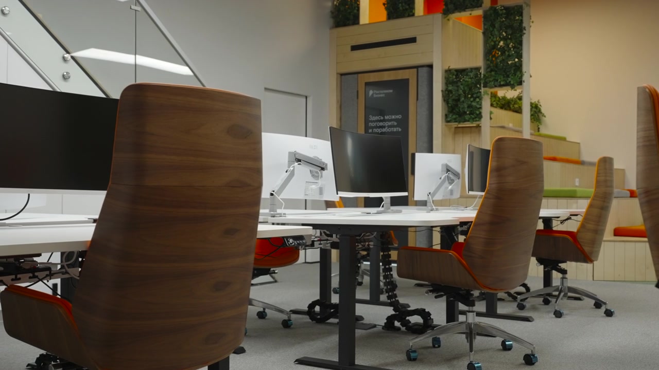 Bright open-plan office space with orange upholstered chairs, desks, computer monitors, partition panels, indoor plants, and warm ambient lighting in a