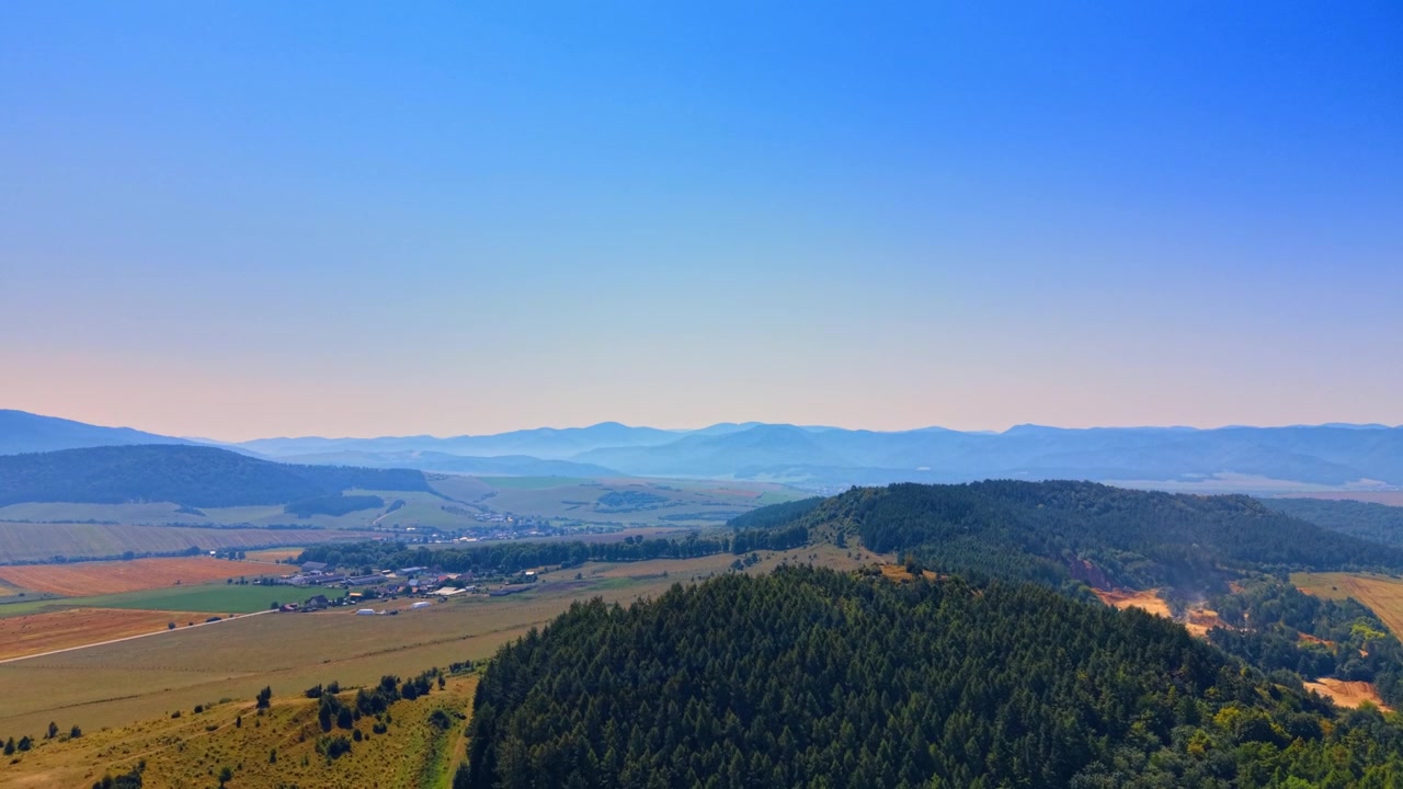Berglandschaft mit bewaldeten Hügeln und Tal. Ein bewaldeter Hügel im Vordergrund mit einem weiten Tal und fernen Bergketten unter klarem Himmel.
