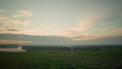 an adult male, in a white shirt, hat, and jeans, stands contemplatively in a grassy field by a tranquil lake at sunset. the serene landscape and warm light create a peaceful, reflective atmosphere