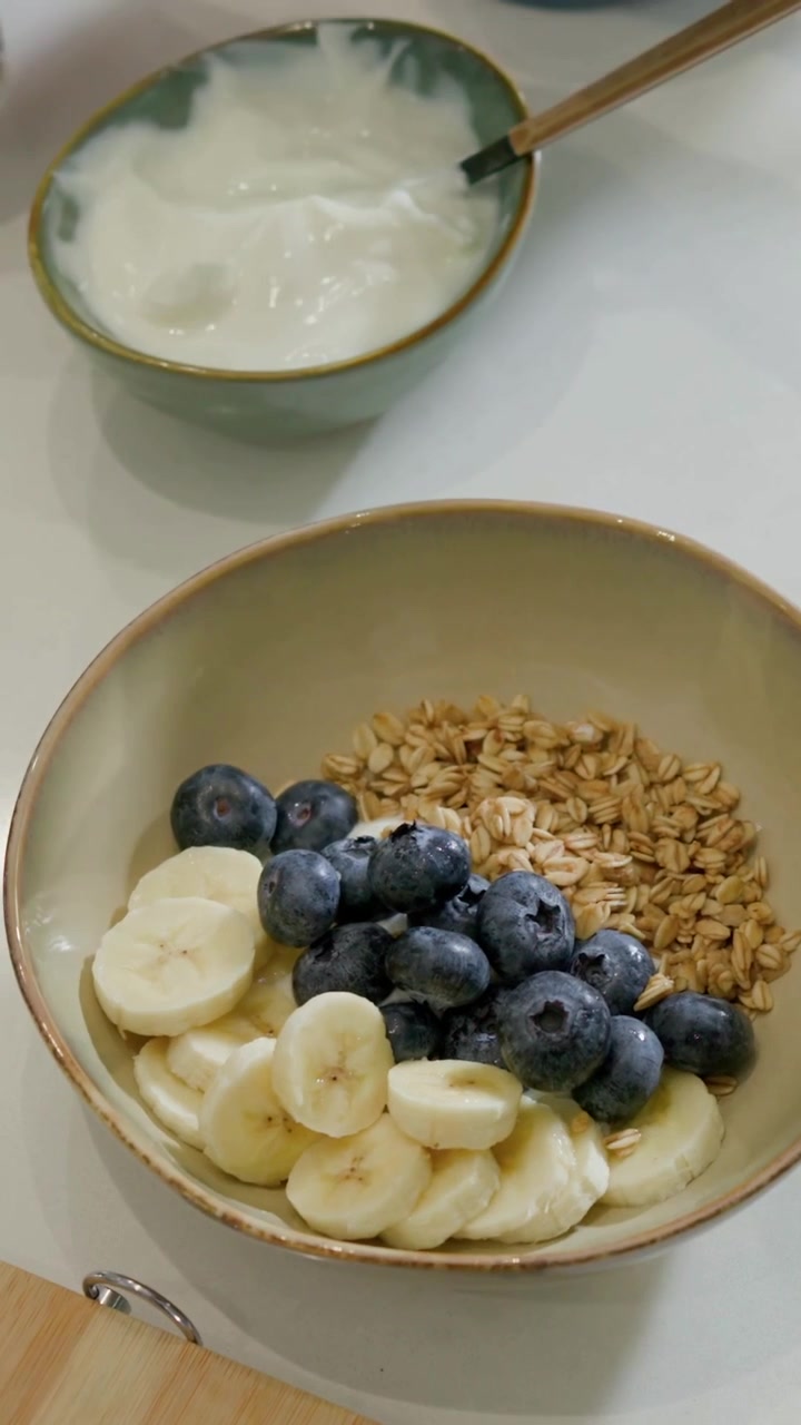 Video verticale: Una mano con polsino blu che mette mirtilli e sistema banane in una ciotola a casa per colazione.