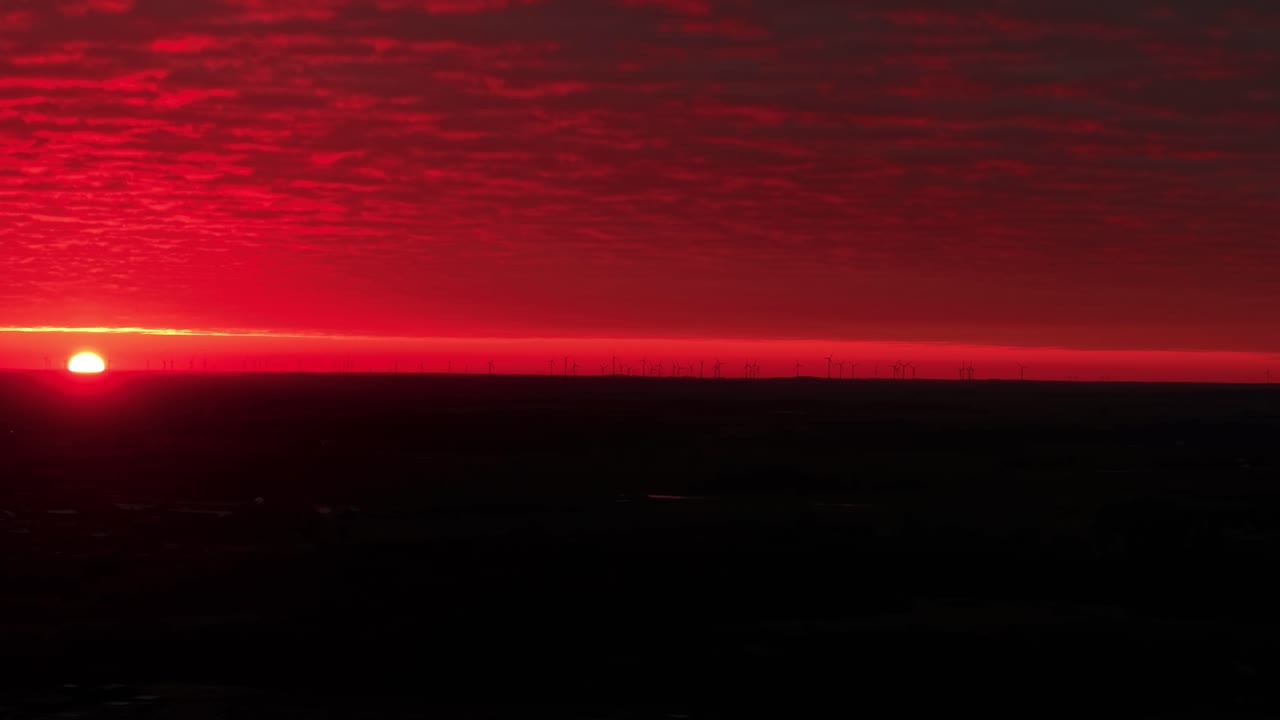 Uma vista aérea de tirar o fôlego captura o pôr do sol sobre um vasto horizonte, pintando o céu em profundos tons de vermelho. A dramática formação de nuvens realça o ambiente sereno e, ao mesmo tempo, impactante.