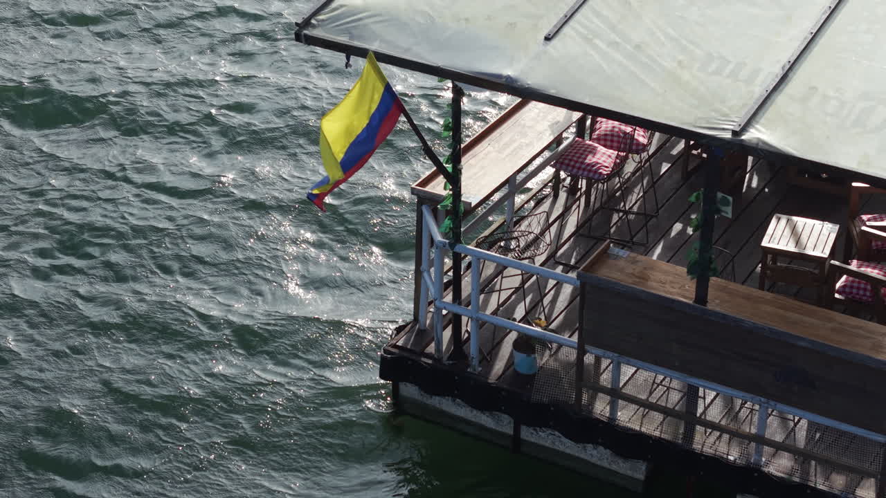 Drone video captures a vibrant floating bar at Lake Calima. Sunlight sparkles on the water, highlighting the Colombian flag. The cozy setting invites relaxation and scenic views