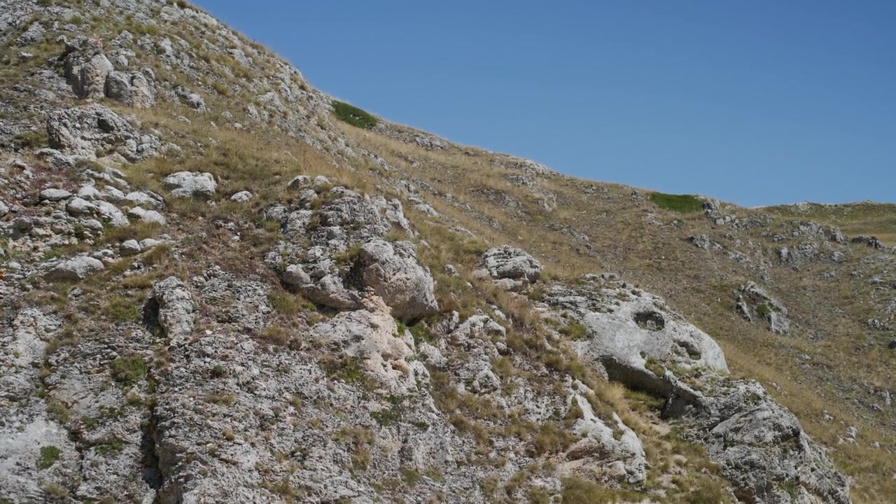 Massi rocciosi su un aspro versante montano sotto un cielo azzurro e limpido. Panoramica a mano libera da sinistra a destra di un ripido pendio alpino con erba secca durante una soleggiata giornata estiva.