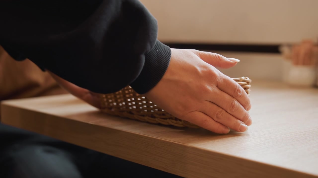 Close-up das mãos de uma pessoa movendo e colocando uma cesta de vime trançado sobre uma mesa de madeira. O fundo inclui um bule de vidro, xícara de cerâmica, iluminação suave, ambiente interno com detalhes de fundo desfocados.