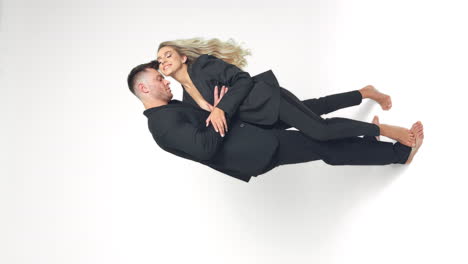 beautiful caucasian couple wearing similar black suits embracing and kissing in studio. man and woman in love at white backdrop. vertical screen.