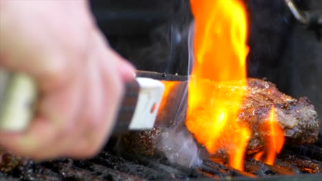 Un-Par-De-Pinzas-Para-Carne-Deslizan-Un-Jugoso-Filete-De-Costilla-Casi-Cocido-En-Una-Parrilla-Y-Lo-Presionan-Mientras-Las-Llamas-Se-Disparan-A-Cámara-Lenta