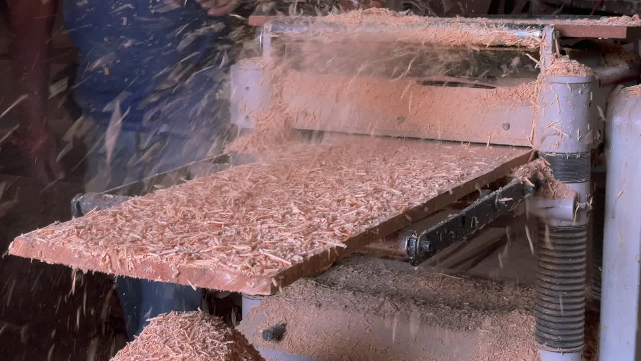 Wood planer machine processes a wooden board with sawdust flying and piling up in a woodworking shop. Static shot