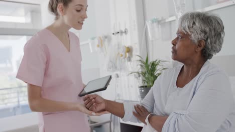 Happy-diverse-female-doctor-using-tablet-talking-with-senior-female-patient-in-bed,-slow-motion