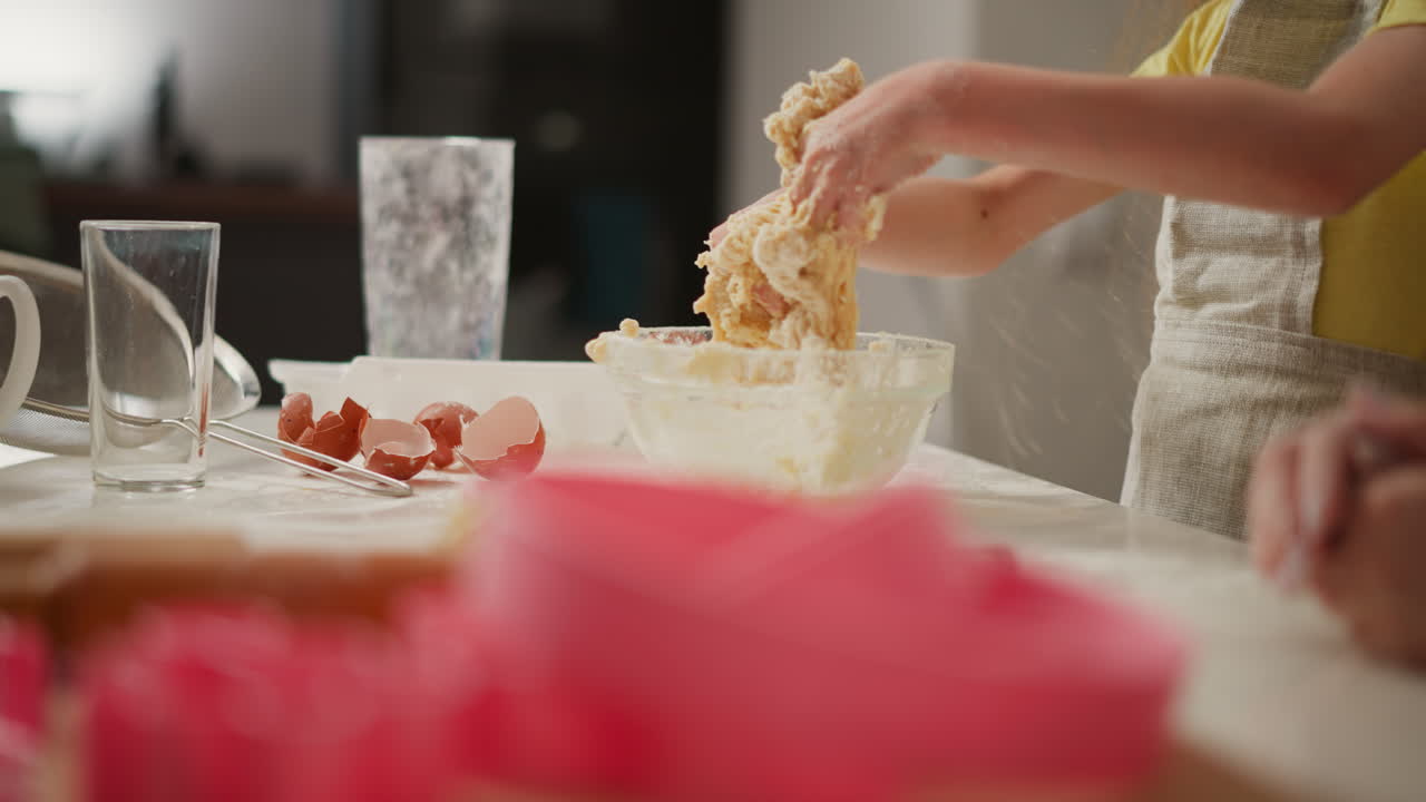 Partial view of person pulling sticky dough off hands above glass bowl during home baking session, with scattered eggshells, glasses, sieve, and utensils in soft kitchen lighting on countertop