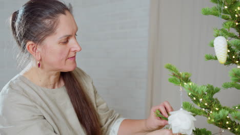 close view of woman fixing white ornament on christmas tree decorated with vibrant green pine branches and glowing lights, festive cozy indoor setting capturing joyful spirit of holiday preparation