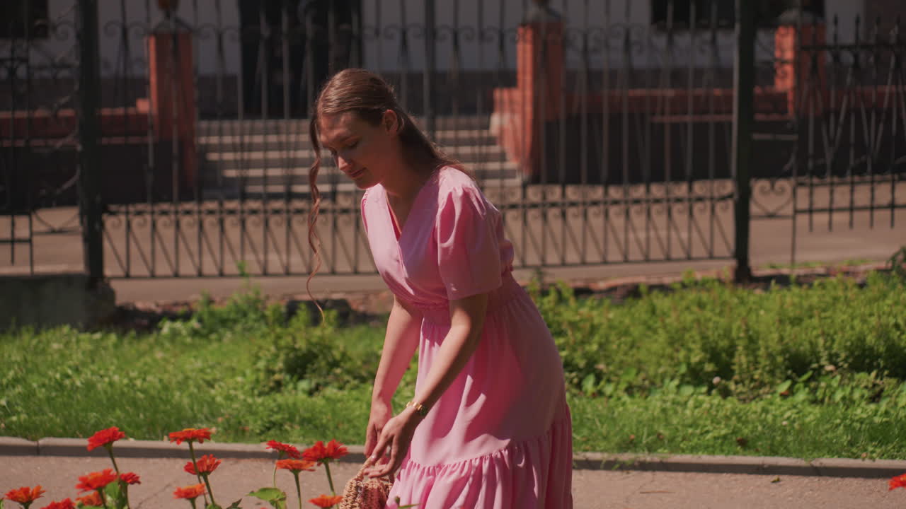 Mujer blanca con vestido rosa inclinada sobre unas zinnias rojas, primer plano oliendo las flores con el pelo trenzado, momento sensorial divertido, curiosidad juvenil, luz brillante sobre los pétalos, intimidad en un sendero de jardín urbano