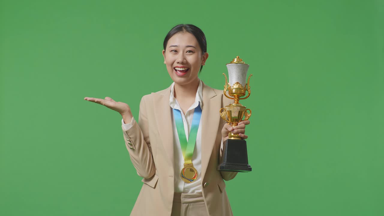 금메달을 가진 슈트를 입은 아시아 사업가 여성이 손에 금 트로피를 들고 미소 짓고 스튜디오의 녹색 화면 배경에 을 가리키고 있습니다.