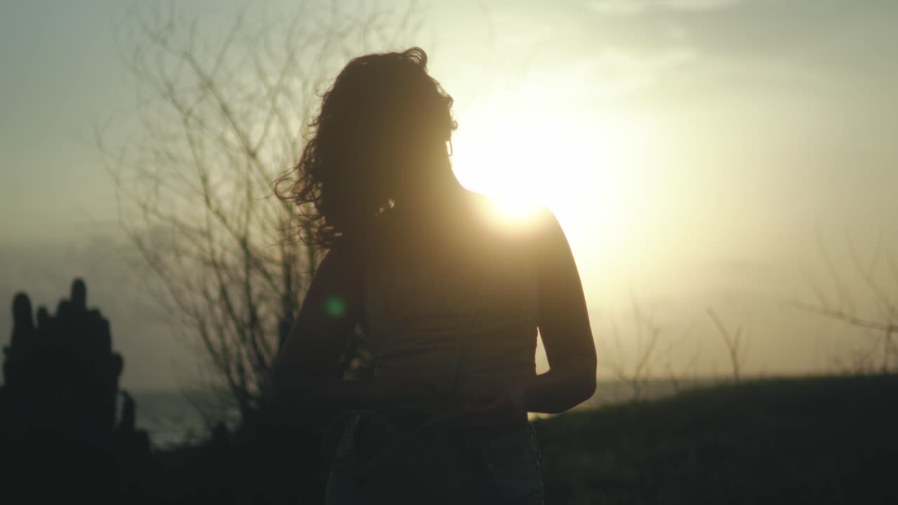 Silhouette di donna con capelli scompigliati dal vento, che si muove all'aperto durante l'ora d'oro, sperimentando la natura, l'ispirazione e il benessere al tramonto.
