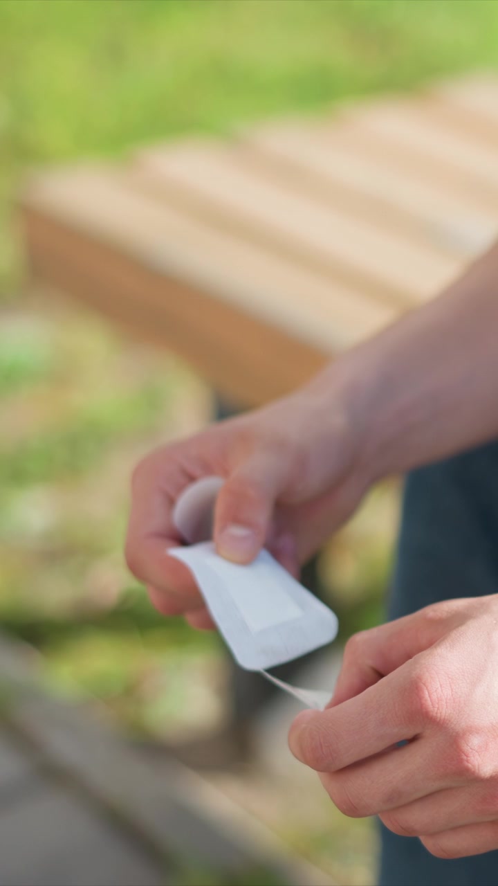Primo piano di mani che strappano un piccolo oggetto di carta bianca in un ambiente esterno con panca di legno ed erba verde. Le mani manipolano materiale sottile mostrando un'azione deliberata contro uno sfondo naturale sfocato