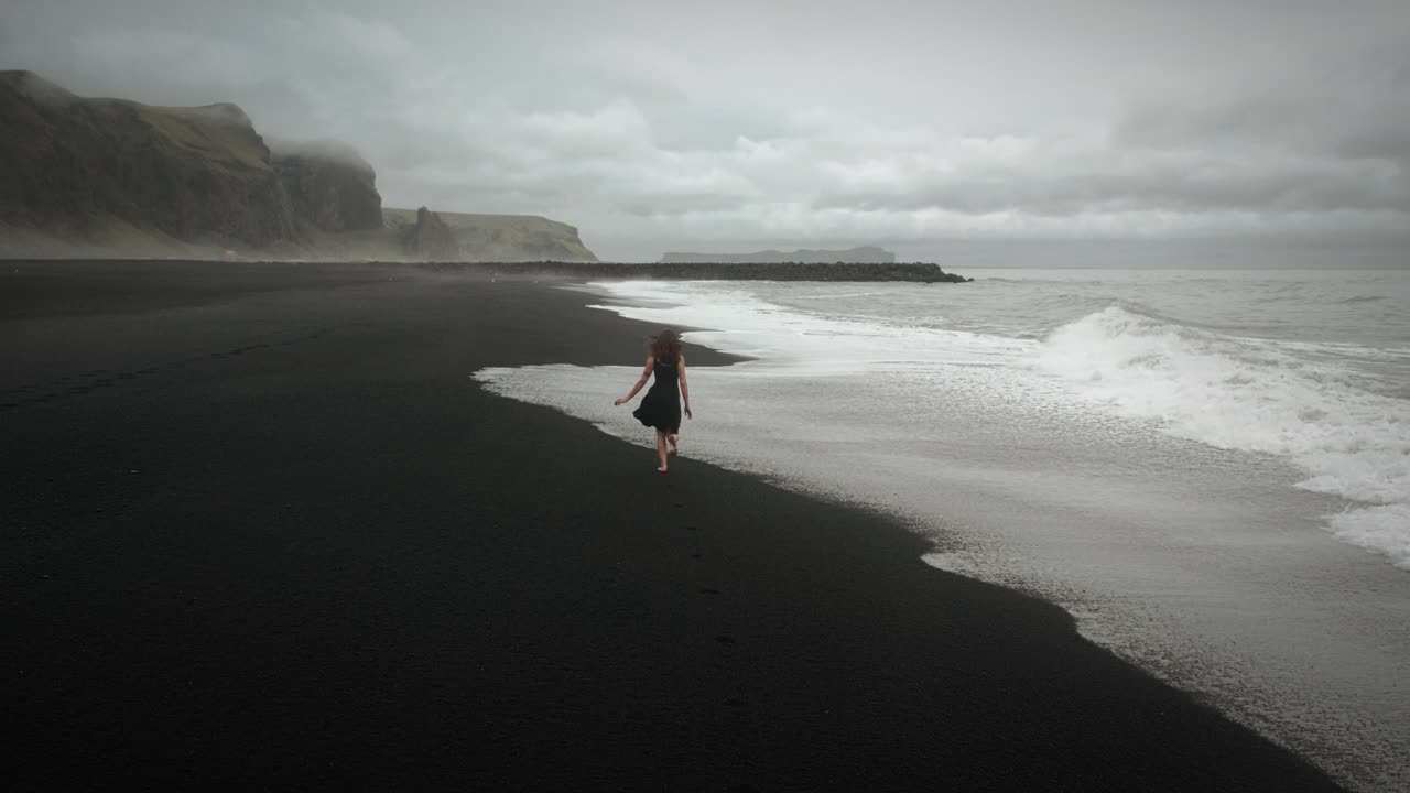 joven hermosa mujer en vestido negro corriendo en la playa de arena negra islandia, disparo de seguimiento