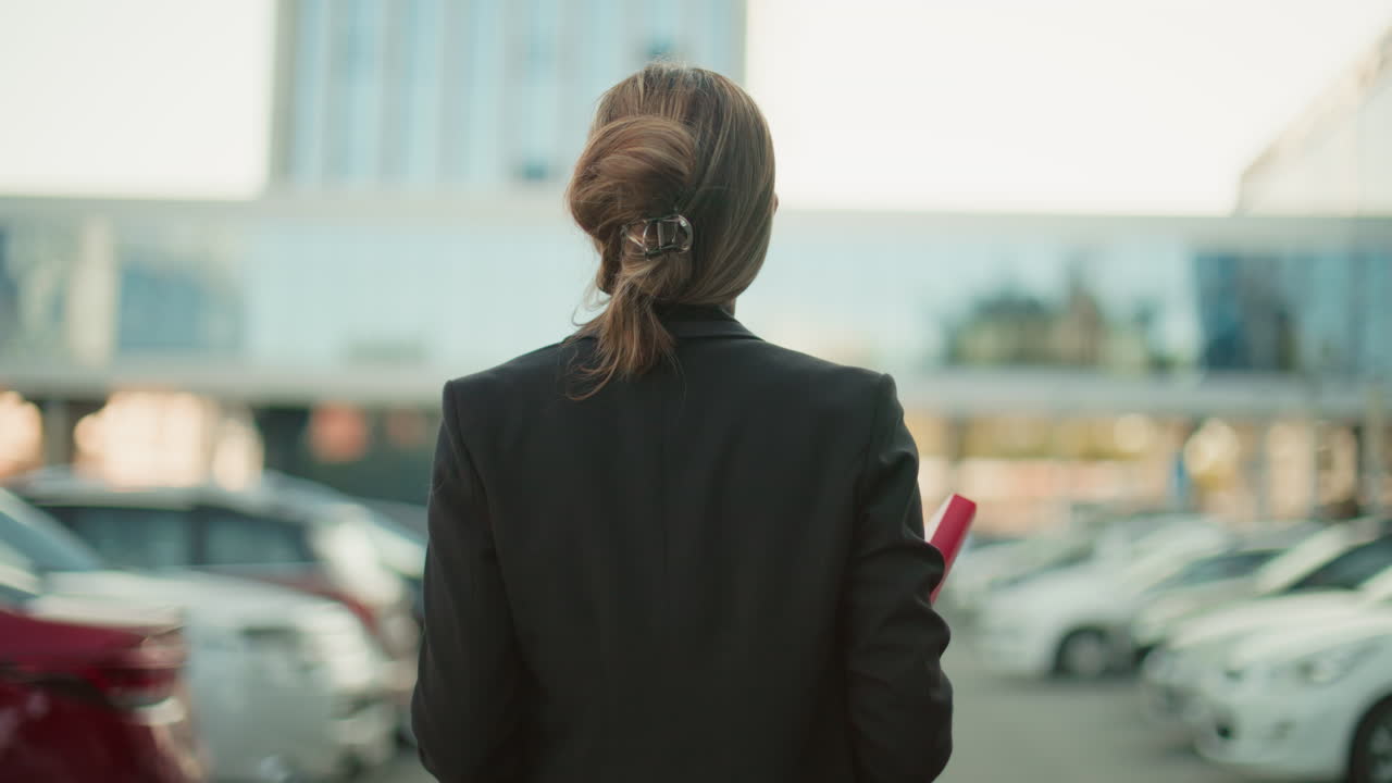 Back view of lady walking with red folder and coffee cup through city parking lot with blurred background of parked cars and glass office building on bright day
