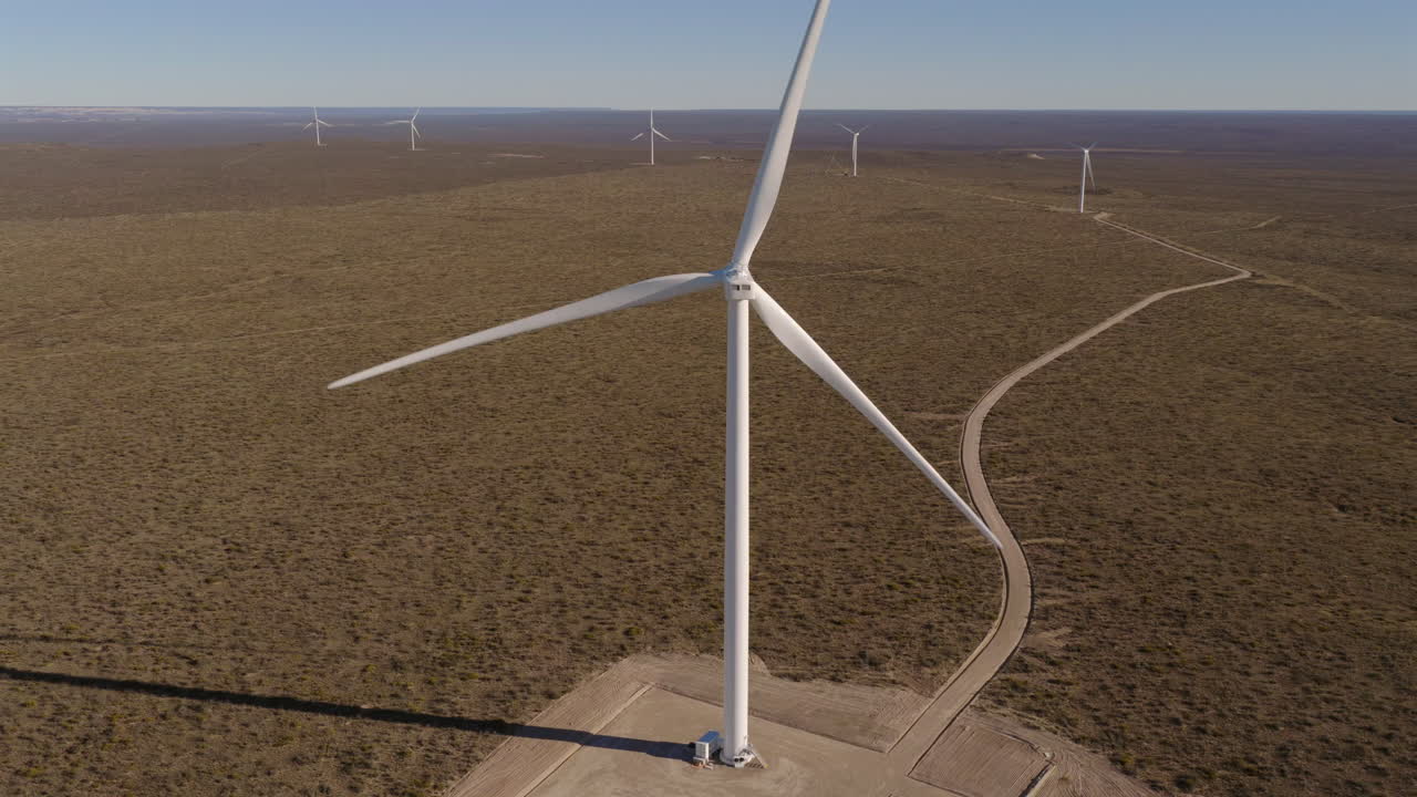 Ascending drone shot looks at single large wind turbine spinning, with others visible in the distant background