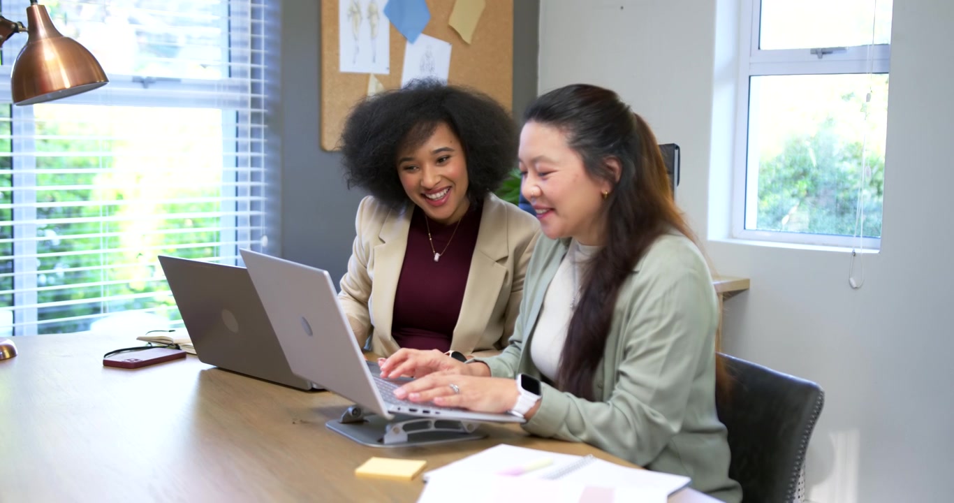 Diverse female coworkers reviewing project draft, right woman typing on raised laptop stand at desk